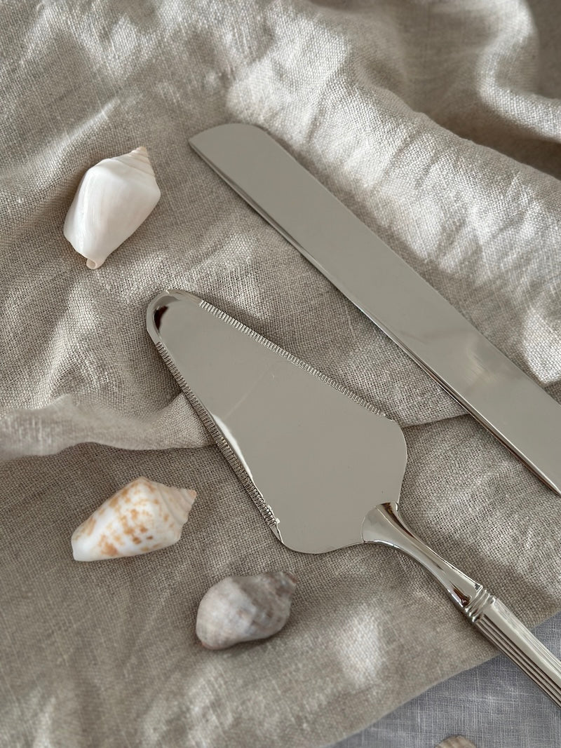 Two metal cake knives on a textured fabric surface with seashells.