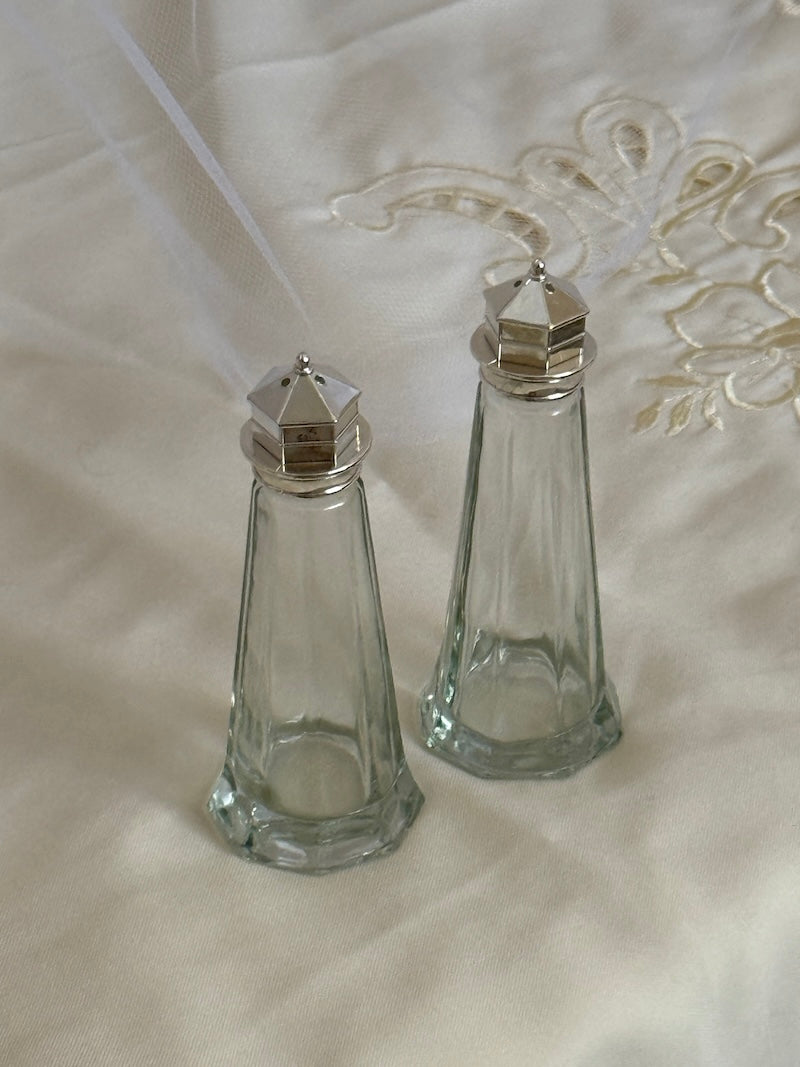 Two clear glass salt and pepper shakers with silver lids on a white fabric background.