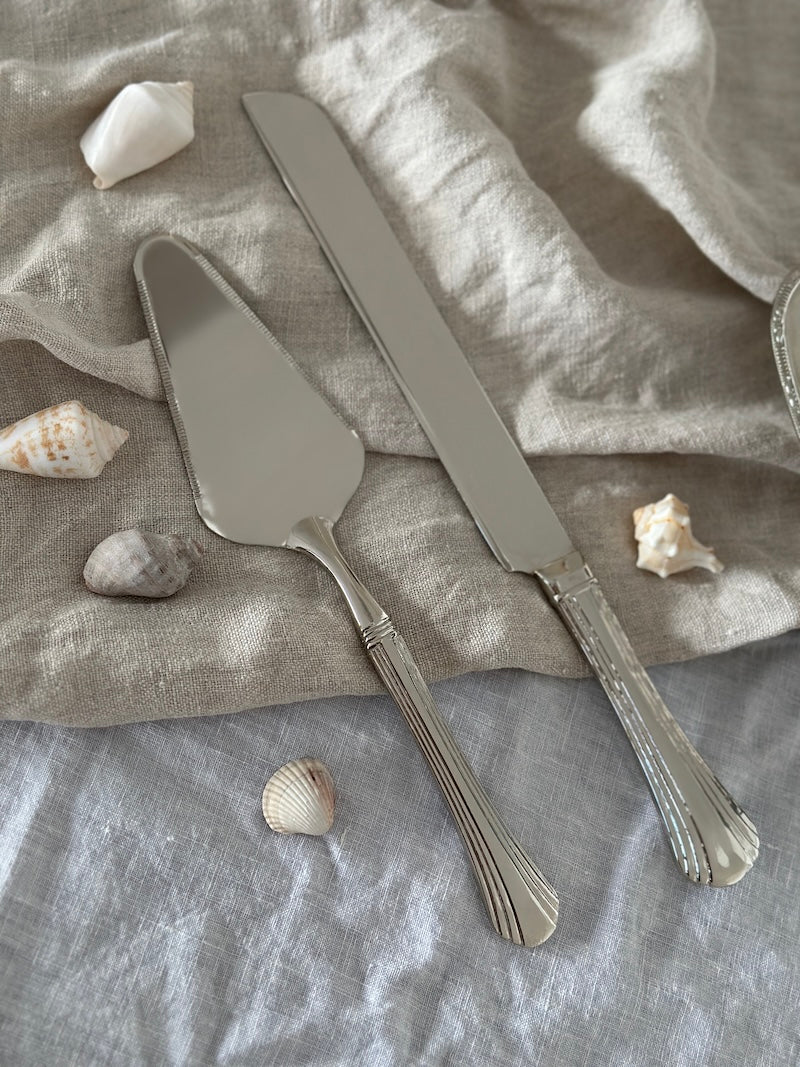Silver cake server and knife set on a textured fabric surface with seashells.